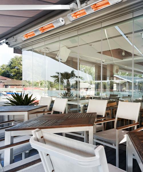 Outdoor restaurant dining area with small wooden diamond-shaped tabled and white chairs with two KMH-30R 3KW Infrared Wall Mounted Outdoor Garden Heaters mounted to the rood above the restaurant's large glass windows