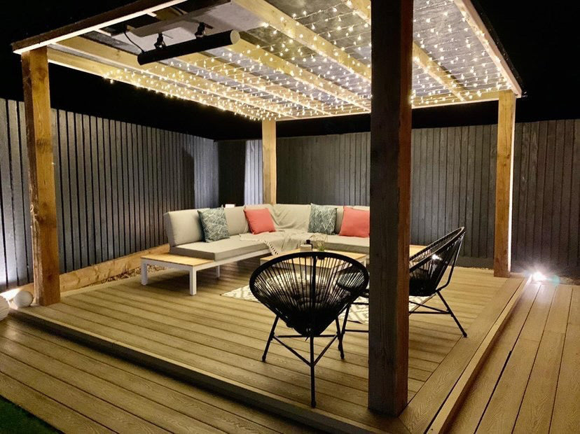 Outdoor wooden patio area lit by fairy lights with a soft grey corner bench,, two metal tub chairs and the KMH-30R 3KW Infrared Wall Mounted Outdoor Garden Heater mounted on a wooden beam