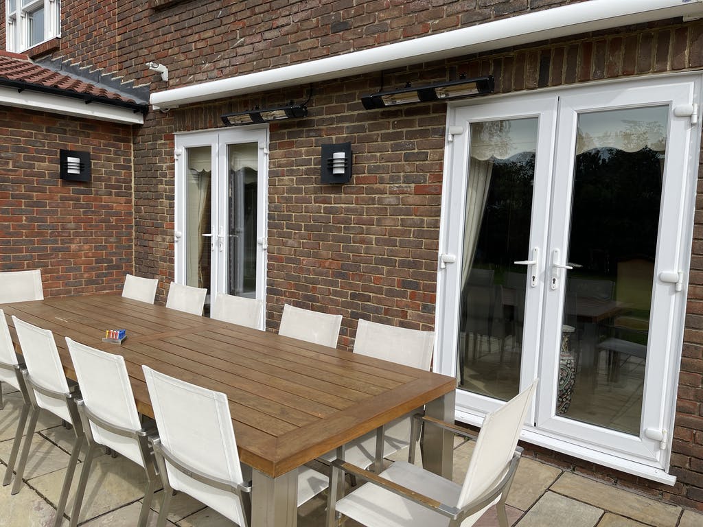 Outdoor stone patio area with a long wooden table and set of ten white chairs with two KMH-30R 3KW Infrared Wall Mounted Outdoor Garden Heaters mounted on the brick house wall