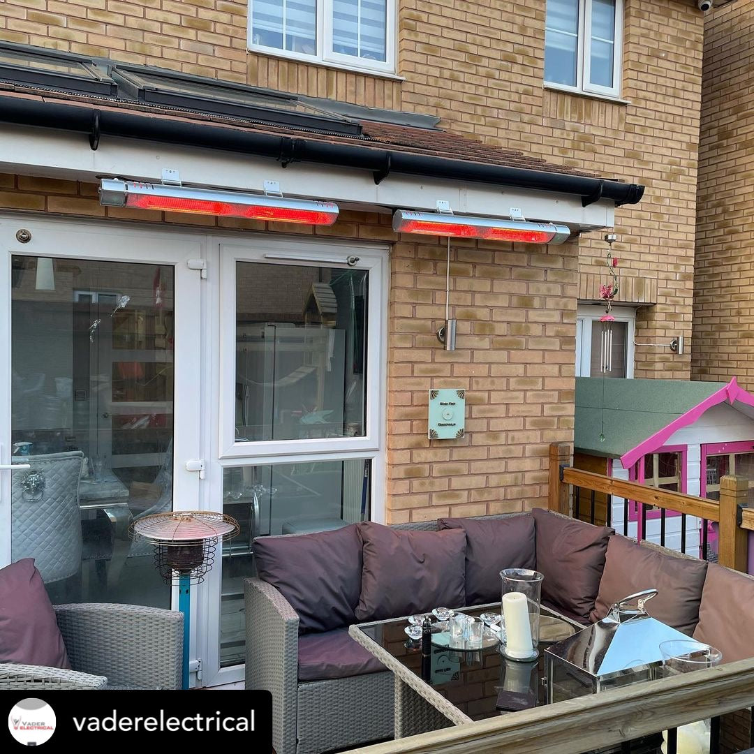 Garden patio area with a grey wicker corner bench with purple pillows, a black glass wicker table and two KMH-30R 3KW Infrared Wall Mounted Outdoor Garden Heaters mounted on the house wall