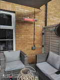 Load image into Gallery viewer, Lifestyle shot of the silver KMH-20 2KW Infrared Outdoor Wall Mounted Garden Heater mounted on the outdoor brick wall of a garden patio house with grey wicker furniture on the deck